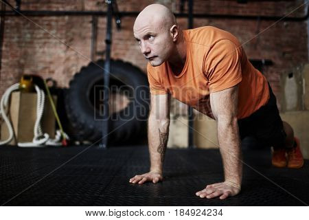 Handsome middle-aged sporty man wrapped up in intensive training: he flexing muscles while doing push-ups in gym, full-length portrait