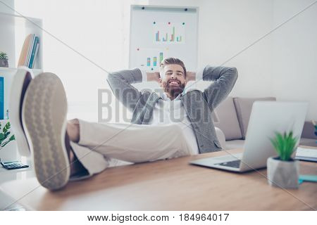 Young Cheerful Businessman Is Resting At A Workplace, With Feet On Top Of The Desk, Smiling