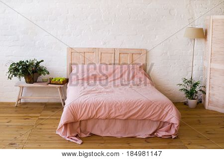 Empty bed with pink pillows and pink cover in the bedroom. White wall wooden floor home flowers with green leaves floor lamp and small table near the bed apples and the book on it. Home interior