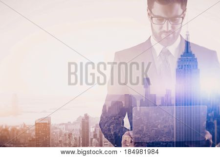Thoughtful young businessman using laptop on creative New York city background. Communication concept. Double exposure