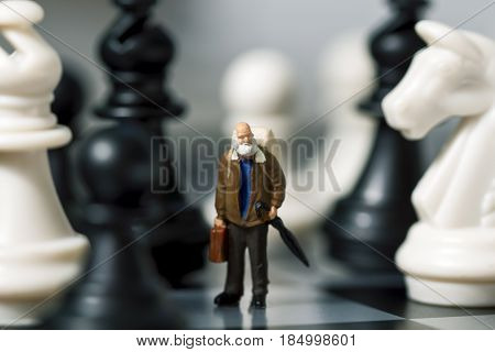 Miniature doll traveler and chess. Old traveler in chessboard. Senior traveller with suitcase. White and black chess figurines. Model learning to play chessmate. Big figures and small man concept