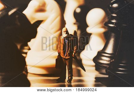Miniature doll and chess vintage toned photo. Old traveler on chessboard. Senior traveller between white and black chess figurines. Model learning to play chessmate. Big figures and small man concept