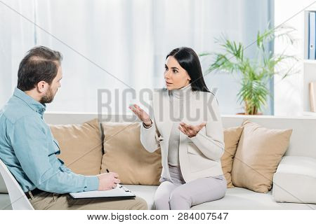 Emotional Female Patient Talking And Looking At Psychotherapist Writing On Clipboard