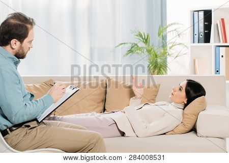 Side View Of Bearded Psychotherapist Writing On Clipboard And Young Patient Lying In Couch