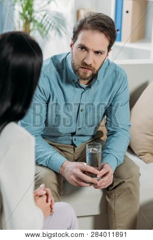 Cropped Shot Of Upset Bearded Man Holding Glass Of Water And Looking At Psychotherapist In Office