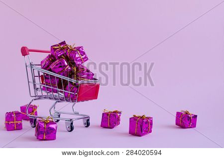 Small Shopping Cart With Presents On Pink Background. Trolley From A Supermarket Full Of Purple Gift