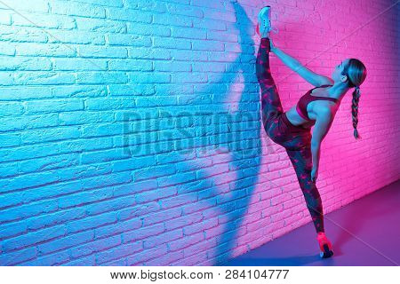 Attractive Young Slim Gymnast Woman In Sports Clothing Stretching On Brick Wall In Neon Lights. Flex