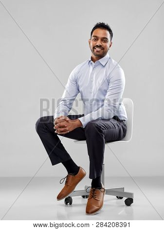 business, furniture and people concept - smiling indian businessman sitting on office chair over grey background