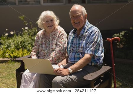 Front view of happy senior couple with laptop sittind on bench and looking at camera on sunny day in garden