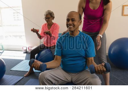 Front view of young female trainer assisting senior man making exercices on exercice ball in fitness studio