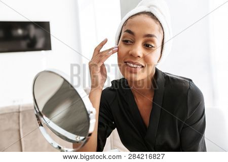 Portrait of young african american woman in housecoat wrapped in white towel applying face cream against mirror