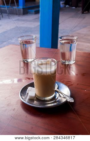 Traditional Moroccan Coffee With Milk In A Simple Glass. Plate, Ornate Spoon And Sugar Cubes. Dirtly