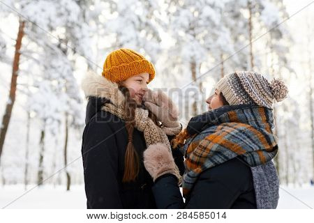 Side View Portrait Of Happy Mother Tying Scarf On Cute Little Girl While Having Fun In Winter Forest