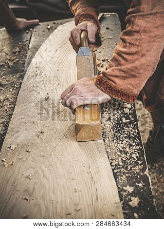 Carpenter In Medieval Cotton Clothes Working With A Wood By Plane. Man Manually Hews A Wooden Board 