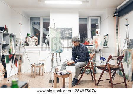 Young Professional Artist Sits On A Chair Near An Easel With A Host In A Cozy Creative Studio And Pa