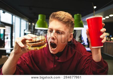 Portrait Of An Emotional Young Man Holding A Glass Of Beverage And A Burger, Looking At A Burger And