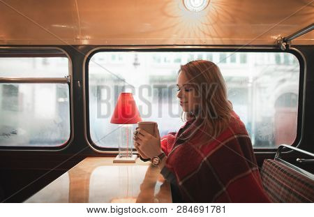 Sweet Lady Sits In A Cozy Cafe At The Window Covered With A Blanket, Holds A Cup Of Coffee In Her Ha