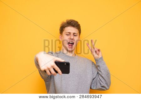 Funny Guy In Casual Clothing Is Standing On A Yellow Background, Take Selfie On A Smartphone And Pos