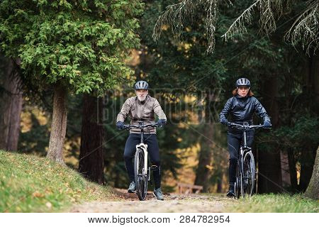 A Senior Couple With Electrobikes Cycling Outdoors On A Road In Park In Autumn.
