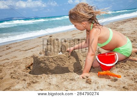 Little girl wearing green swimsuitbuilding a sand cactle on a sandy beach in Crete, Greece. Family summer vacation concept