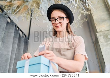 Young woman in eyeglasses and hat tying up knot and making bow on to pof giftbox while binding it with silk ribbon