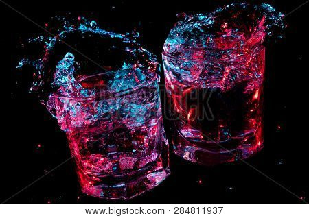 Two Rocks Glasses Clinking Together And Spilling Liquid Under Colorful Lights Isolated On A Black Ba