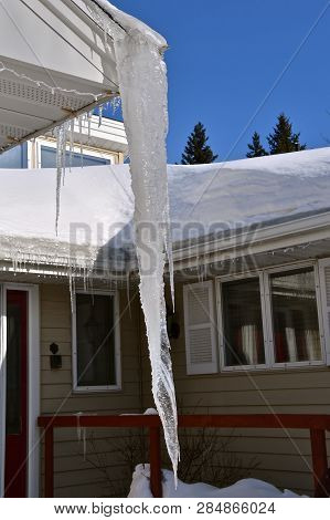 A Huge Icicle Hangs From The Eavestrough Of A House With A Roof Loaded With Snow