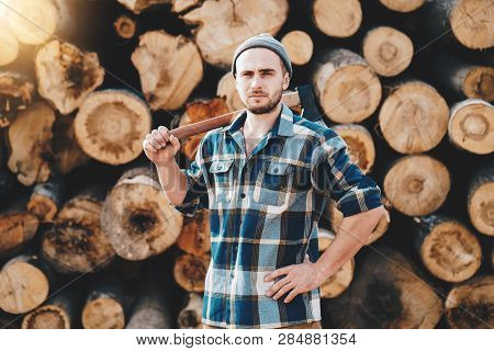 Strong Bearded Lumberman Wearing Plaid Shirt Holds Axe On His Shoulder On Background Of Sawmill