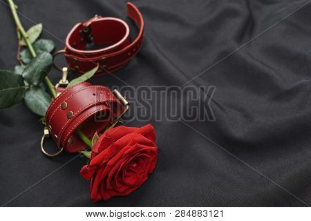 Erotic Toys. Top View Of Red Leather Handcuffs And Rose Against Of Black Silk Fabric.