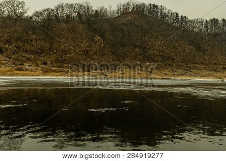 Landscape Of Partially Frozen River On Winter Day With Hilly Tree Lined Shore In Background.