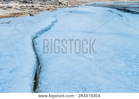 Closeup Of Long Crack In Ice Sheet On Frozen River With Sandy Rocky Shoreline.