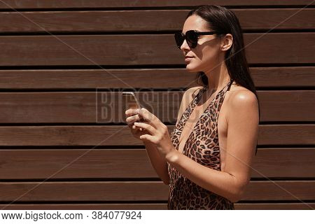 Woman In Bikini And Black Sunglasses Holding Smart Phone In Hands, Looking In Distance, Posing Isola