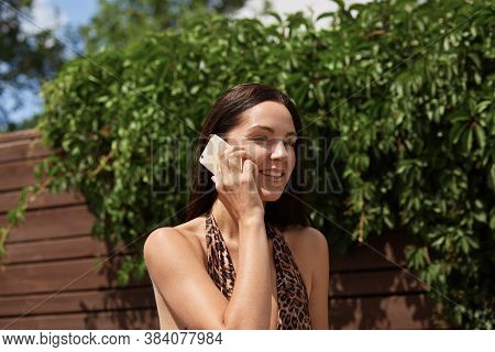 Calm Smiling Female In Swimsuit With Leopard Print Standing Near Green Trees And Speaking On Smart P
