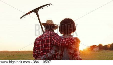 Rear On African American Couple Of Farmers Walking In Countryside With Pitchfork Outside And Hugging
