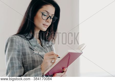 Sideways Shot Of Hard Working Executive Woman In Spectacles, Formal Checkered Suit, Writes Down Info