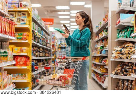A Young Beautiful Caucasian Woman Reads The Information On The Label Using Her Mobile Phone. The Con