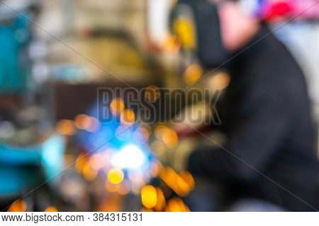 Blurred Background Of Electric Welding Process. Worker In Welding Mask. Yellow Sparks Fly To The Sid