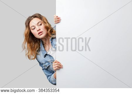 Young Woman Peeking Out From Behind A White Wall. Mockup Of The Announcement Or Presentation.
