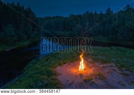 Midsummer Ligo Celebration. Drone View Of Huge Fire On The River Shore. Scenic Landscape Of Forest A