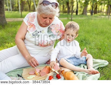 Happy Grandmother And Grandson Eating Sandwiches On Picnic At Summer Park. Boy Relaxing On Blanket O