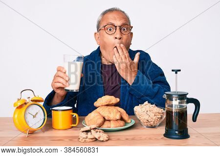 Senior handsome man with gray hair sitting on the table drinking a glass milk for breakfast covering mouth with hand, shocked and afraid for mistake. surprised expression 