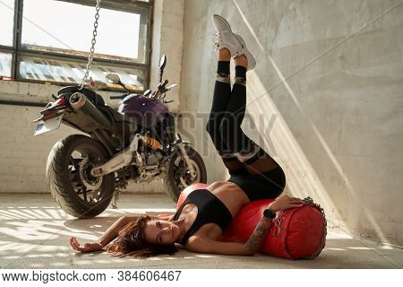 Woman With Multiple Hobbies Posing Near Her Motorcycle And Posing Leaning On Punching Bag In Trainin