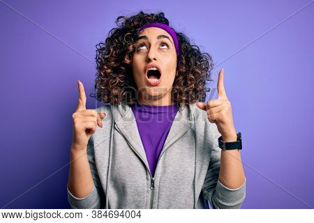 Beautiful curly arab sportswoman doing sport wearing sportswear over purple background amazed and surprised looking up and pointing with fingers and raised arms.