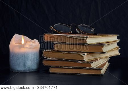 A Stack Of Old Books, A Burning Candle And Glasses On A Dark Background