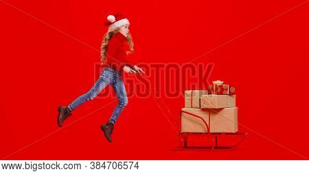Merry Christmas and Happy New Year! Happy girl child in Christmas hat pushing the sled with gifts. Studio portrait over festive red background.