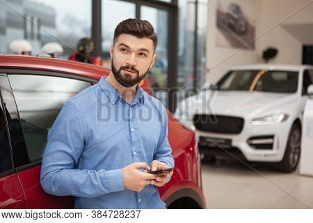 Young Handsome Man Looking Away Thoughfully While Using Smart Phone At Car Dealership, Copy Space