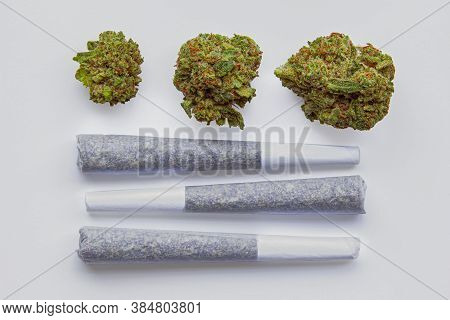 Three Pre-rolls Of Cannabis Joints With 3 Dried Cannabis Flowers On The Side.