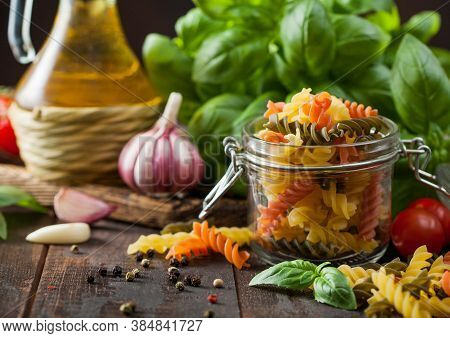 Glass Jar Of Fresh Raw Tricolor Fusilli Pasta With Oil And Garlic, Basil Plant And Tomatoes On Woode