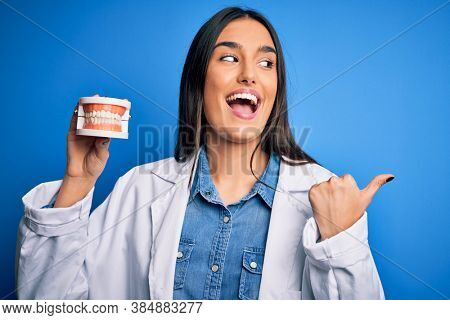 Young beautiful brunette dentist woman wearing coat holding plastic teeth prosthesis pointing and showing with thumb up to the side with happy face smiling