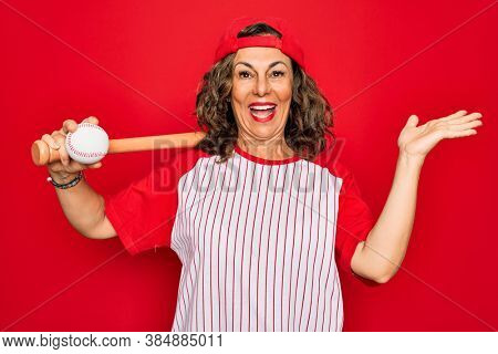 Middle age senior woman wearing baseball equiment, bat and ball over red isolated background very happy and excited, winner expression celebrating victory screaming with big smile and raised hands